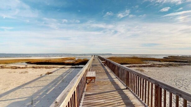 En träpromenad som leder till en strand med sand och gräsbevuxna områden.