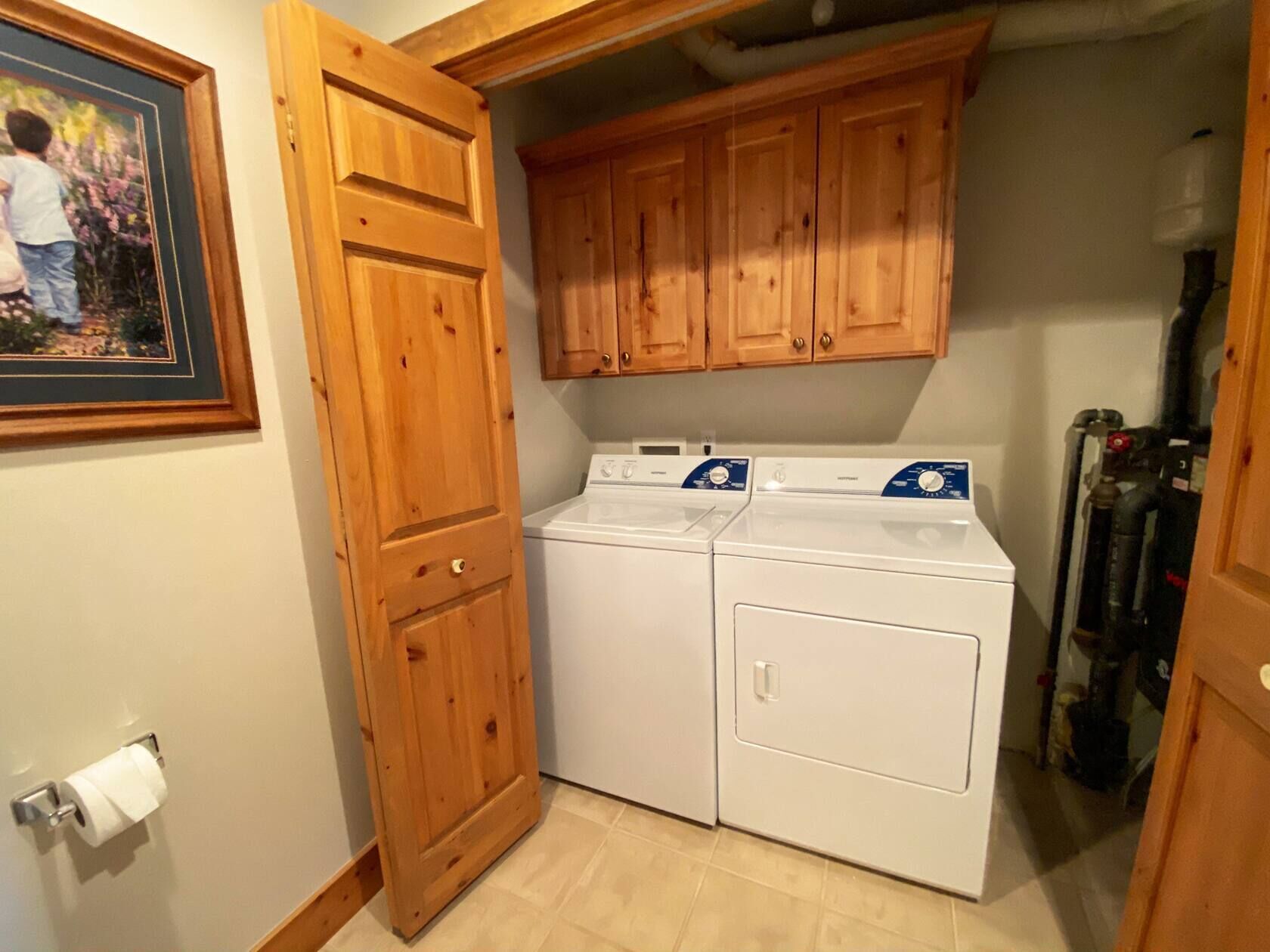 Une buanderie équipée de deux machines à laver et de deux sèche-linge à chargement frontal, tous deux de couleur blanche, de meubles en bois et d’un tableau encadré accroché au mur.