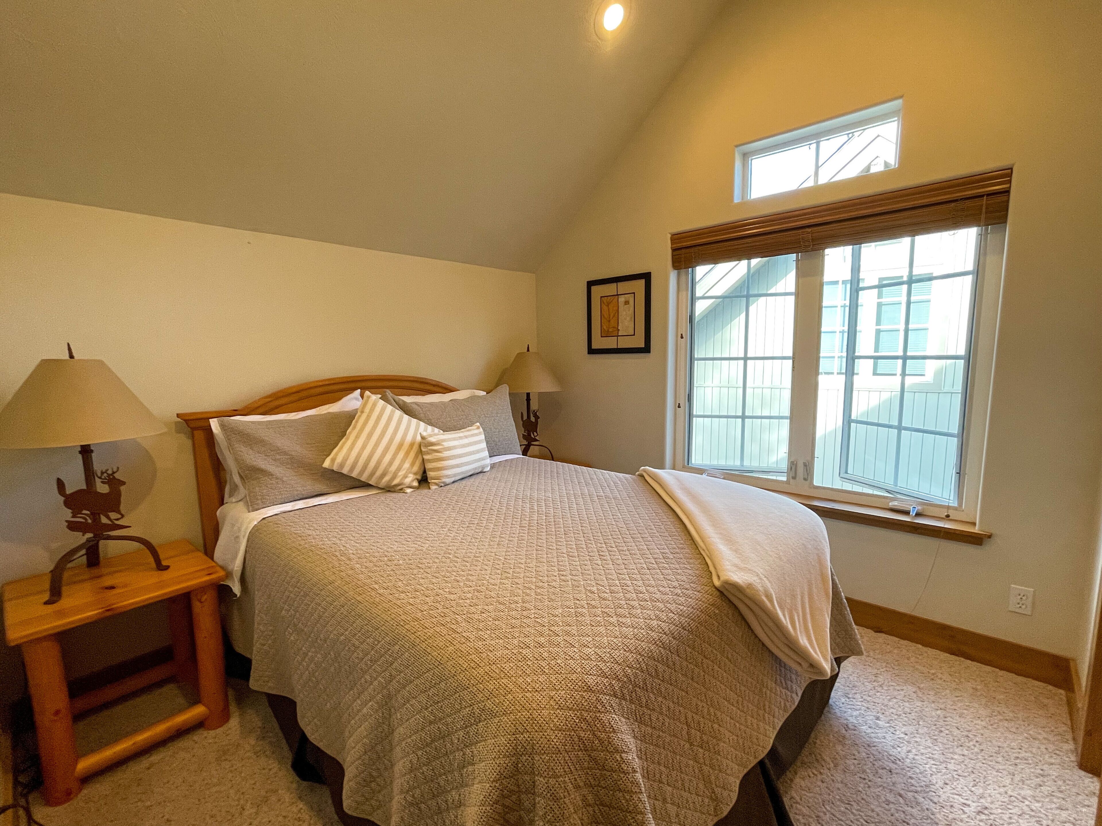 A bedroom with a bed, two bedside lamps, a small wooden nightstand, a window with blinds, and a framed picture on the wall.