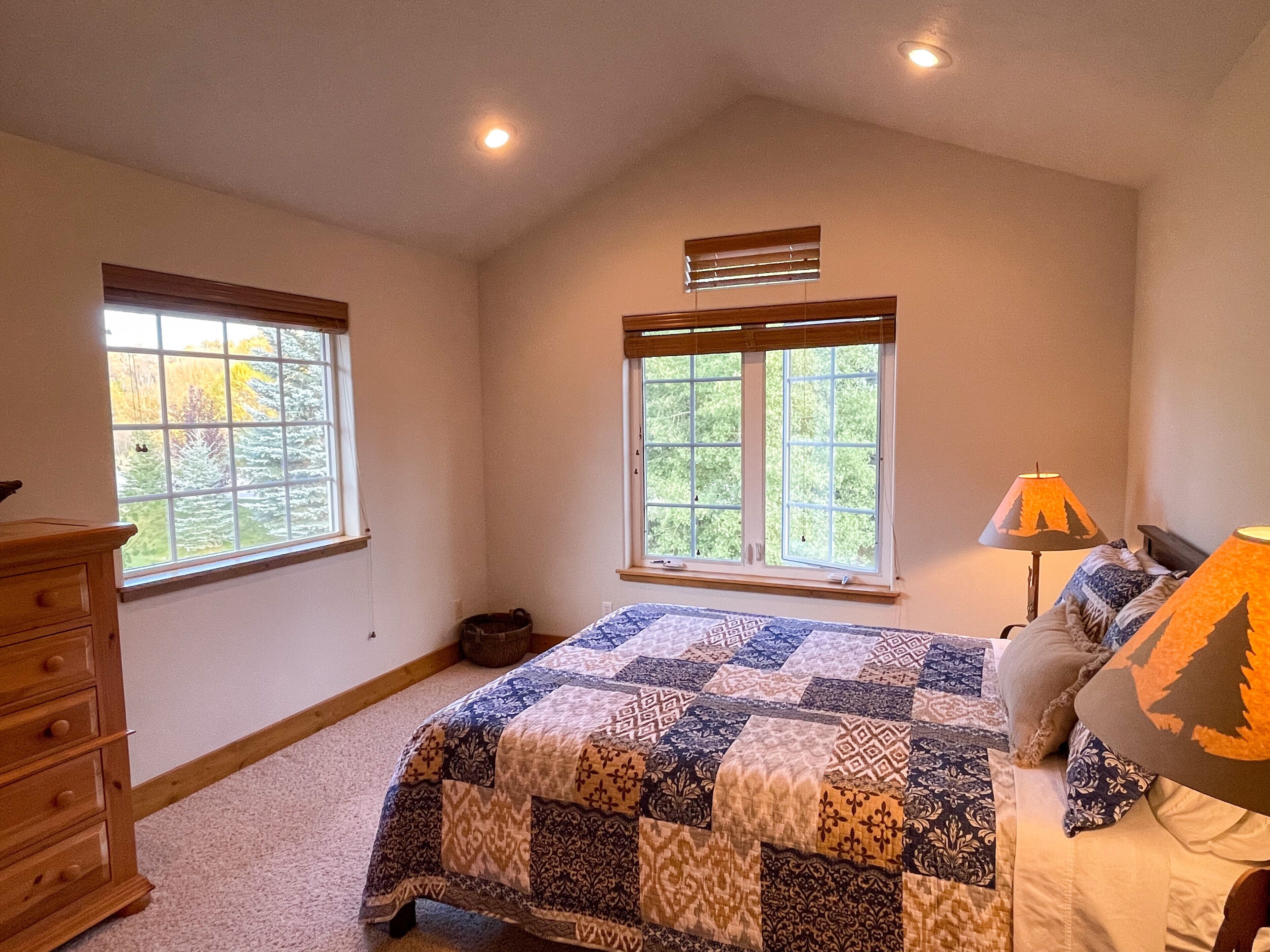 A bedroom with a large bed, two windows, a dresser, and a lamp.