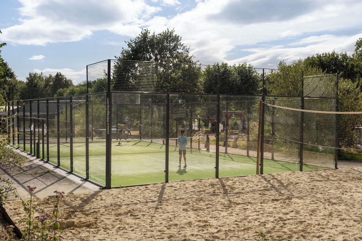 A tennis court enclosed by a net and surrounded by a fence, with a sandy area in front.