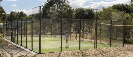 A tennis court enclosed by a net and surrounded by a fence, with a sandy area in front.