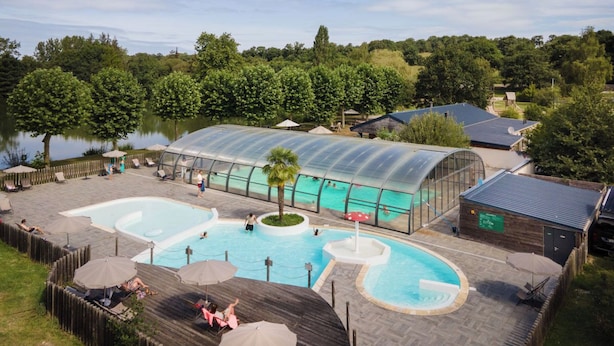 A swimming pool area with two pools, a covered seating area, and a view of a lake. - Huttopia Baie du Mont Saint Michel (Baguer-Pican)