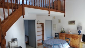 A cozy living room with a wooden staircase, a dining area with a table and chairs, and a kitchen area with a counter and cabinets. - CAPBRETON – Residence LE BOSQUET AUX ECUREUILS. Villa with shared swimming pool. (CAPBRETON)
