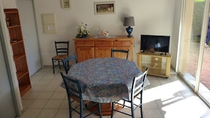 A dining area with a round table, chairs, a wooden cabinet, a television, and a window with a view of greenery. - CAPBRETON – Residence LE BOSQUET AUX ECUREUILS. Villa with shared swimming pool. (CAPBRETON)