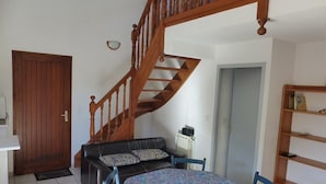 A room with a wooden staircase, a black leather sofa, a round table with a patterned tablecloth, and a wooden bookshelf. - CAPBRETON – Residence LE BOSQUET AUX ECUREUILS. Villa with shared swimming pool. (CAPBRETON)