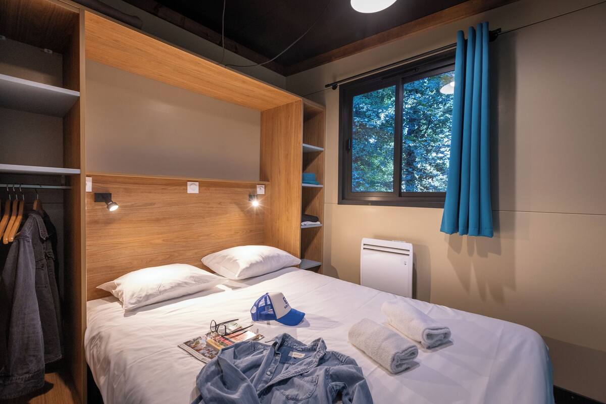 A compact hotel room with a bed, a wardrobe, a window with curtains, and a small desk with a lamp.