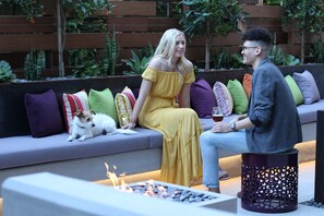 A couple sitting on a modern outdoor sofa with a dog, surrounded by greenery and a fire feature.
