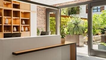 A reception area with a wooden shelving unit displaying vases, a reception desk, and a view of an outdoor garden with potted plants.