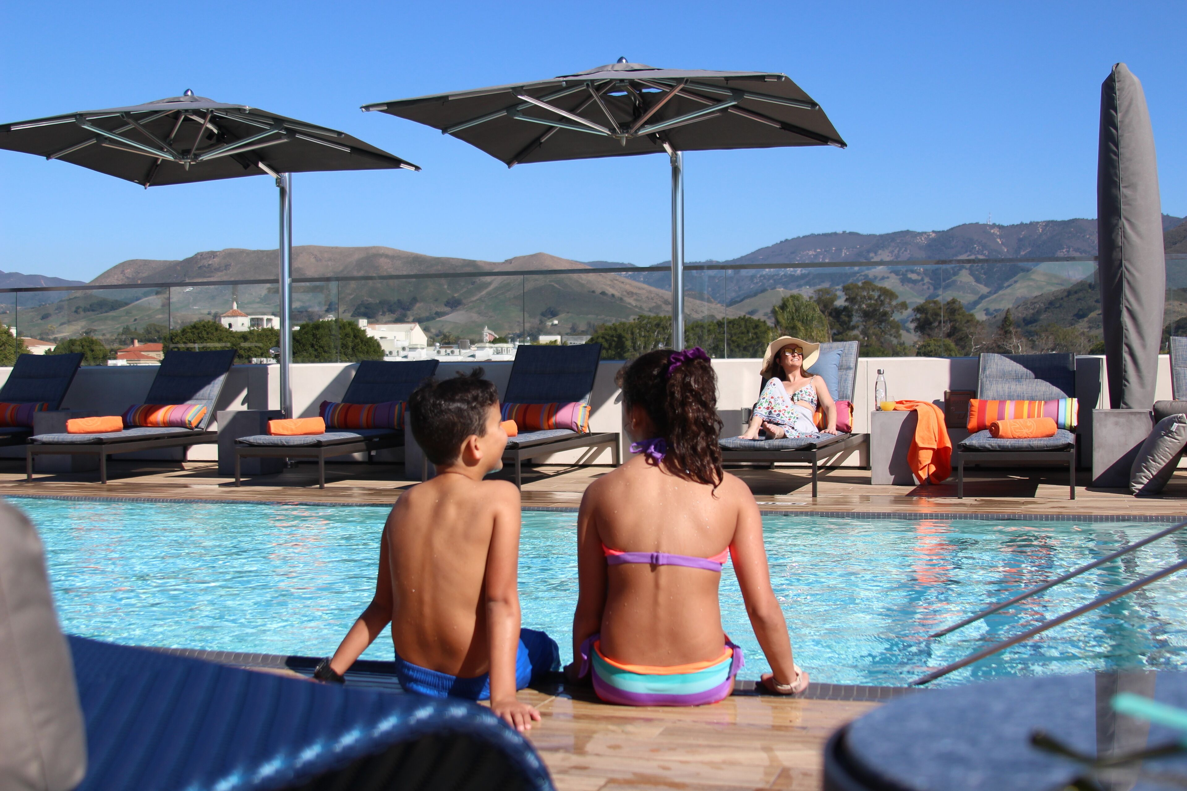 Uma área de piscina com espreguiçadeiras e guarda-sóis, vista desobstruída das montanhas ao fundo, e uma pessoa sentada à beira da piscina.