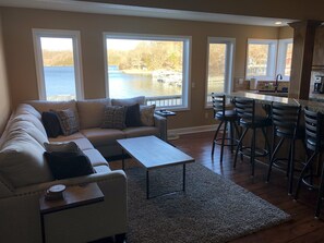 A living room with a sofa, coffee table, and bar stools. Large windows offer views of a lake and dock. - Fully Remodeled Lakefront Home with Beautiful Views of Lake of the Ozarks! (Camdenton)