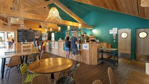 Een café interieur met houten balken in het plafond, een centrale houten tafel en een balie met personeel.