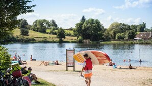 Een strand aan een meer met mensen die zwemmen en zonnebaden, een badmeester met een surfplank, en een bord in de achtergrond.