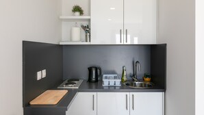 Pieni, käytännöllinen keittiö, jossa on valkoiset kaapit, musta työtaso, vedenkeitin, leikkuulauta, pesuallas ja pieni kasvi hyllyllä.