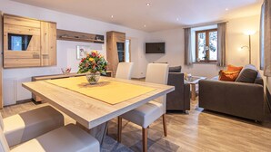 A modern living room with a sofa, a dining table with chairs, a TV mounted on the wall, and a window with curtains. - Vacation apartment Watzmannblick Maria Gern (Berchtesgaden)