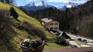 A two-story house with a red roof, surrounded by a forested hillside and snow-capped mountains in the background. - Vacation apartment Watzmannblick Maria Gern (Berchtesgaden)
