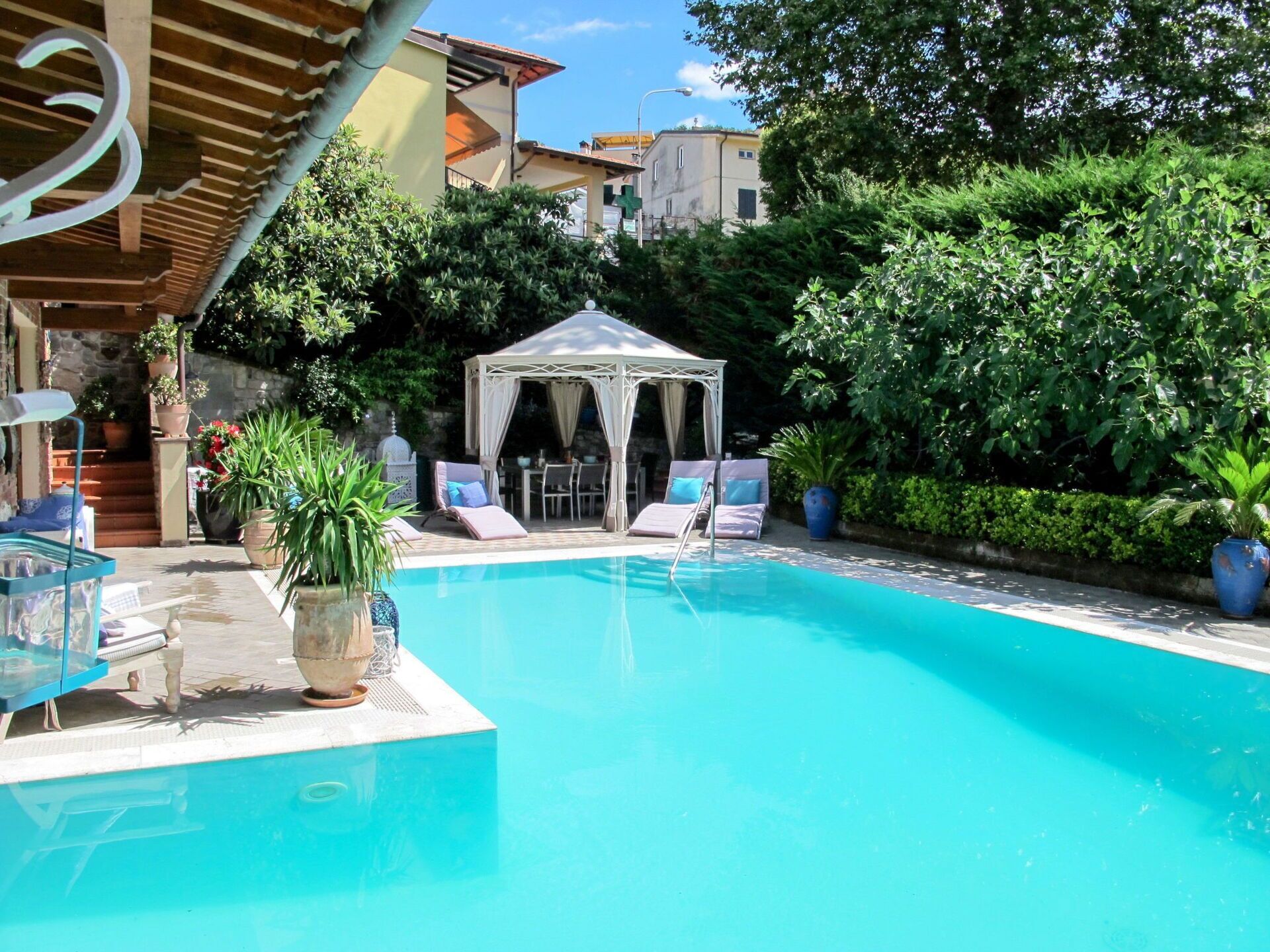 Piscina com espreguiçadeiras e um gazebo, cercada por vegetação e edifícios.