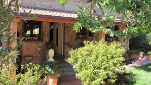 Une maison en pierre avec un toit en tuiles, un porche à l’avant et un jardin agrémenté de diverses plantes ainsi que d’un espace aménagé pour s’asseoir.