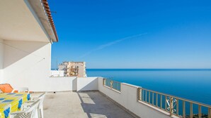 A balcony with a view of the sea, a table with chairs, and a building in the background.