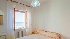 A bedroom with a bed, a window with curtains, a radiator, a nightstand with a lamp, and a view of the sea.