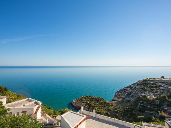 A coastal view with a clear sky, a calm sea, and a rocky landscape with buildings.