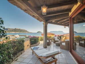 Relax in a serene outdoor space with a clear view of the sea and distant hills, surrounded by lush greenery and a low stone wall.