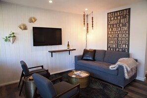 A modern living room with a grey sofa, a flat-screen TV, a wooden shelf, and a decorative wall sign listing various locations.