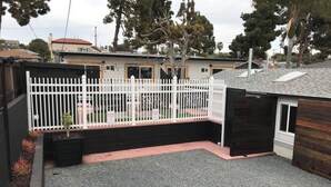 A fenced backyard with gravel, a wooden fence, and a house with a red roof. - Kauai-Inspired California Beach Retreat, Unit 1 (Oceanside)
