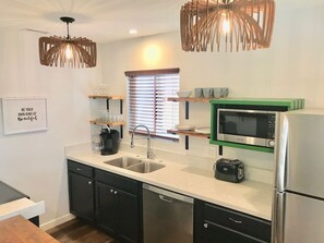 A modern kitchen with a double sink, a microwave, a toaster, and a refrigerator. There are wooden shelves above the sink and a wall-mounted light fixture. - Kauai-Inspired California Beach Retreat, Unit 1 (Oceanside)