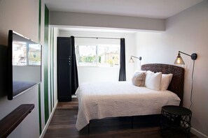 Un dormitorio con cama, lámpara de noche, mesita y ventana con cortinas.