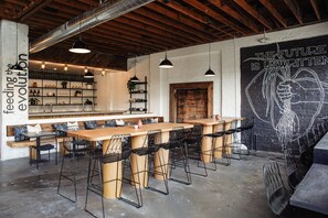Een moderne bar met houten tafels en zwarte metalen stoelen, een bakstenen muur met muurschildering en zichtbare leidingen aan het plafond.