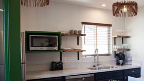 Una cocina moderna con microondas integrado, cafetera y lavavajillas. Hay estantes de madera sobre la encimera y una ventana con persianas.
