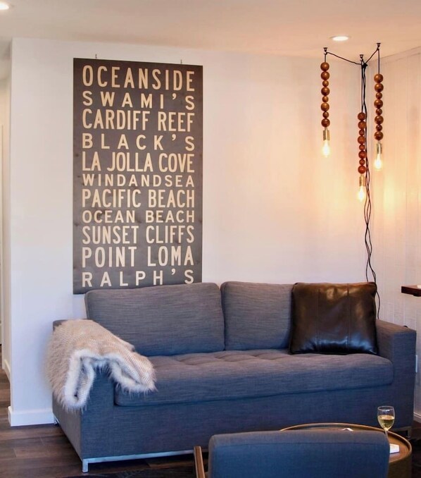 A modern living room with a grey sofa, a decorative wall hanging with place names, and a unique pendant light fixture.