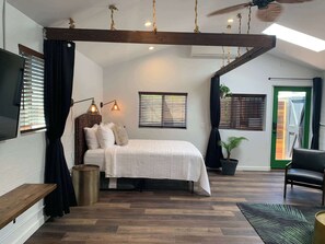 A bedroom with a bed, bedside lamps, a ceiling fan, a window with blinds, a potted plant, and a door with a green frame. - Kauai-Inspired California Beach Retreat, Unit 3 (Oceanside)