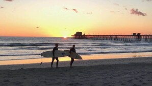 A beach scene with two people carrying surfboards, a pier in the background, and the sun setting over the ocean.