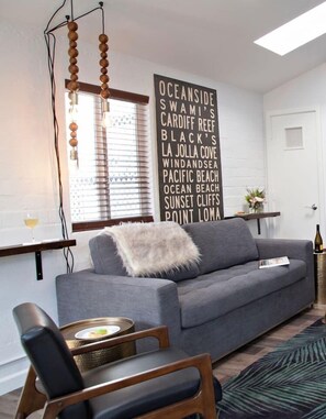 Un salon moderne avec un canapé gris, une chaise en bois, une table avec un vase et un mur orné d’une liste de destinations.