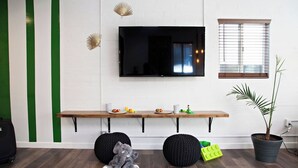 A room with a TV mounted on the wall, a wooden bench with small items on it, two round black stools, a potted plant, and a suitcase on the floor.