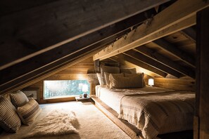 Ein gemütliches Schlafzimmer im Dachgeschoss mit Holzbalkendecke, einem großen Fenster, einem Bett mit Kissen und einem kleinen Nachttisch.