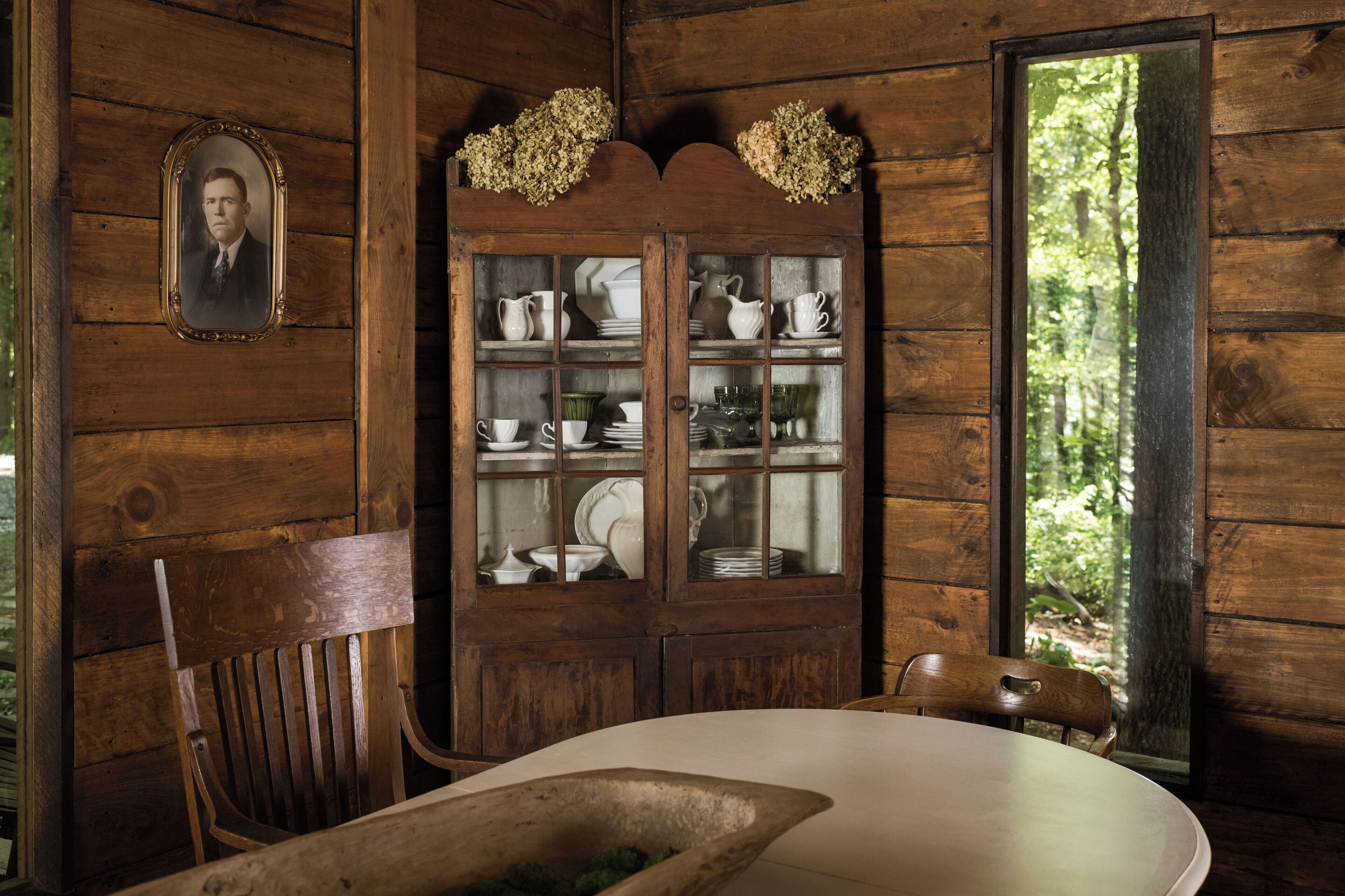 Un interior rústico de madera con una mesa de comedor, sillas, un aparador con vajilla y una fotografía enmarcada en la pared.