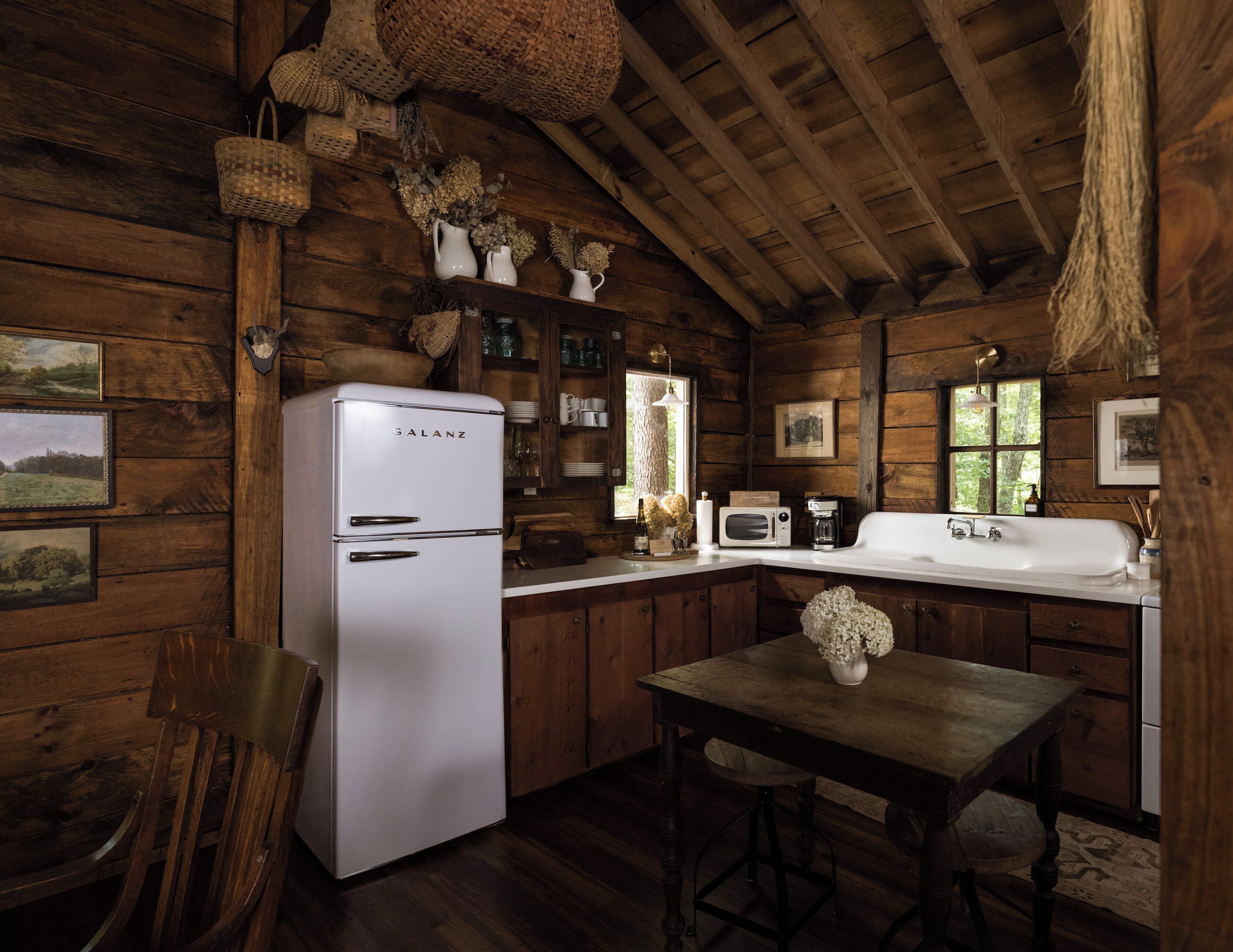 Una cocina rústica con un refrigerador Galanz blanco, armarios de madera, una mesa de comedor de madera y una cesta colgante.