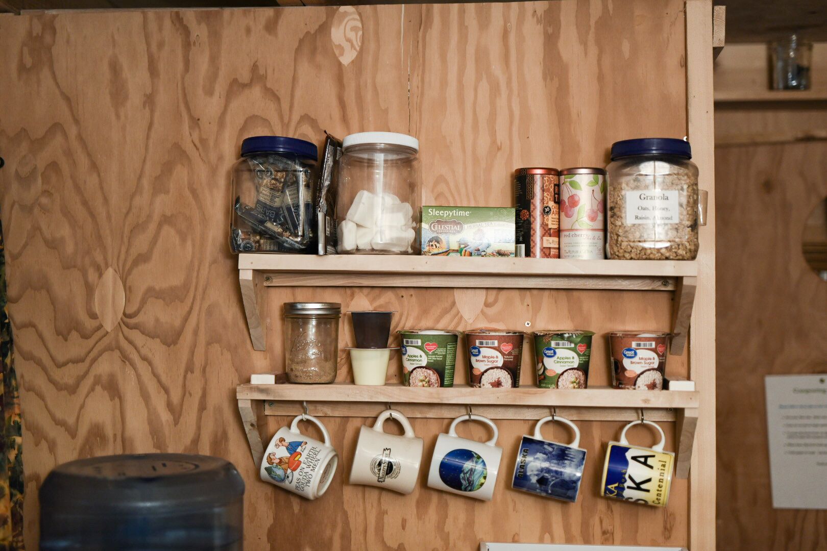 A wooden shelf with jars of granola and other food items, and mugs hanging below.