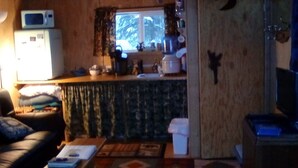 Una sala de estar acogedora con un sofá, una mesita de centro y un televisor. Hay una zona de cocina con un microondas y una ventana con cortinas.