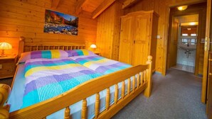 Une chambre à coucher aménagée dans une cabane en bois, avec un grand lit, des murs en bois, un tableau représentant un paysage et une salle de bain visible à travers une porte ouverte.