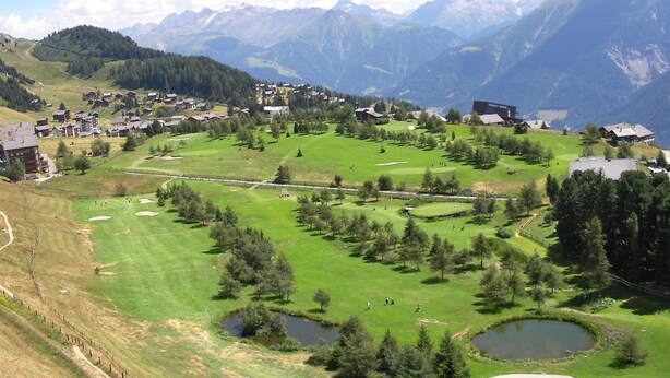 Un parcours de golf comportant plusieurs trous, entouré de bâtiments résidentiels et avec des montagnes en arrière-plan.