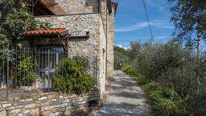 En bygning i sten med et tag af fliser, et hegn i metal og en smal sti, der fører gennem et skovområde.