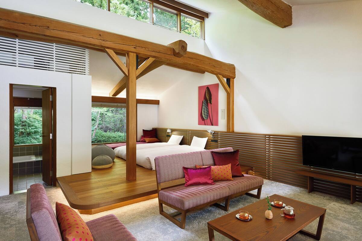 A spacious room with a large bed, a sofa, a coffee table, and a TV. There is a window with a view of trees and a wooden ceiling structure.