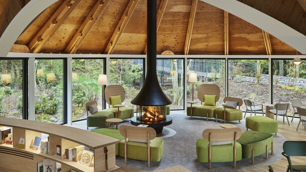 Een gezellig leeshoekje met een open haard, groene gestoffeerde stoelen en een houten boekenkast.