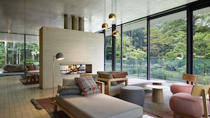 Een moderne woonkamer met een bank, stoelen, een salontafel en een open haard. Grote ramen bieden uitzicht op een bebost gebied.