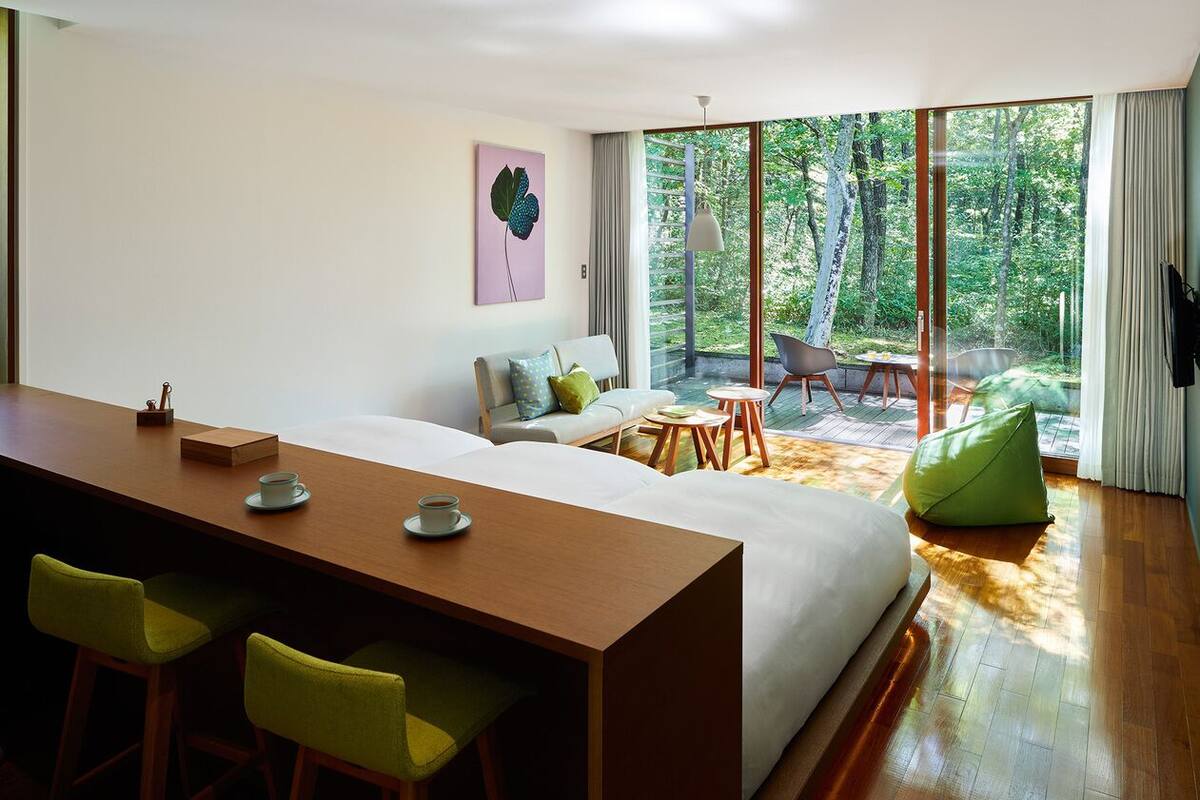 A modern hotel room with a large bed, a wooden desk, two chairs, a small table, and a view of a forest through large windows.