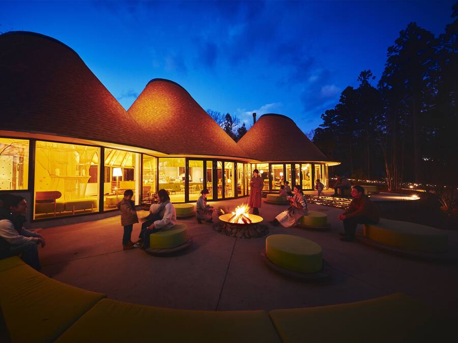 A modern building with a unique curved roof design, large glass windows, and outdoor seating areas with a central fire pit.